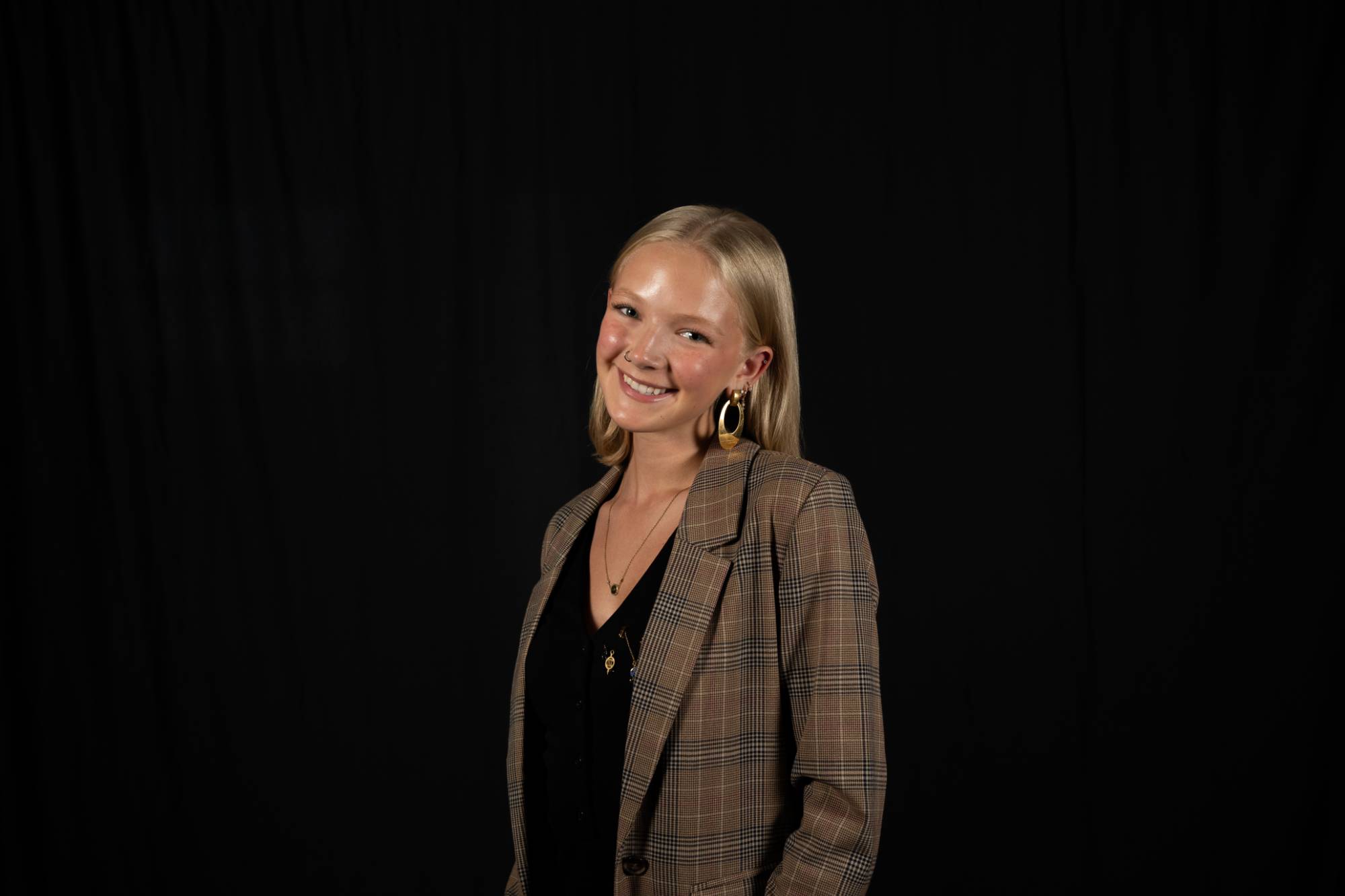 Young woman with blonde hair and plaid jacket smiling at the camera with a black background